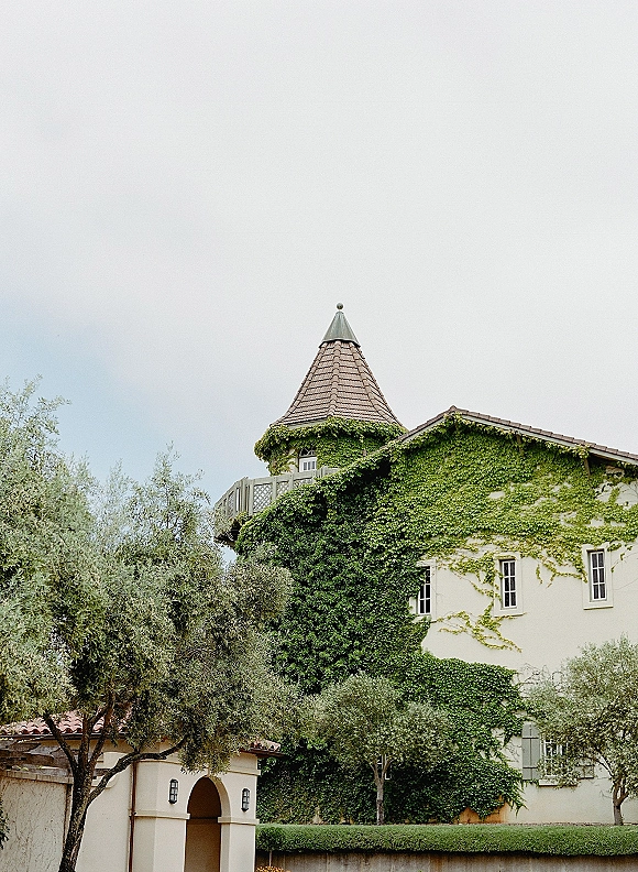 Wedding venue exterior with ivy-covered walls, turret roof, and arched entryway, framed by olive trees and hedges under an overcast sky