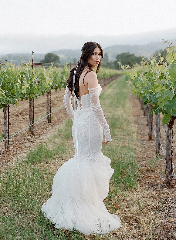 Bridal portrait of a bride in a vineyard wearing an off-the-shoulder lace gown with corset back, long gloves, and tulle train under overcast sky