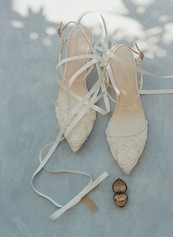 Bridal shoes flatlay with lace wedding heels tied in white ribbon, beside a heart ring box and gold hair comb on sunlit stone surface