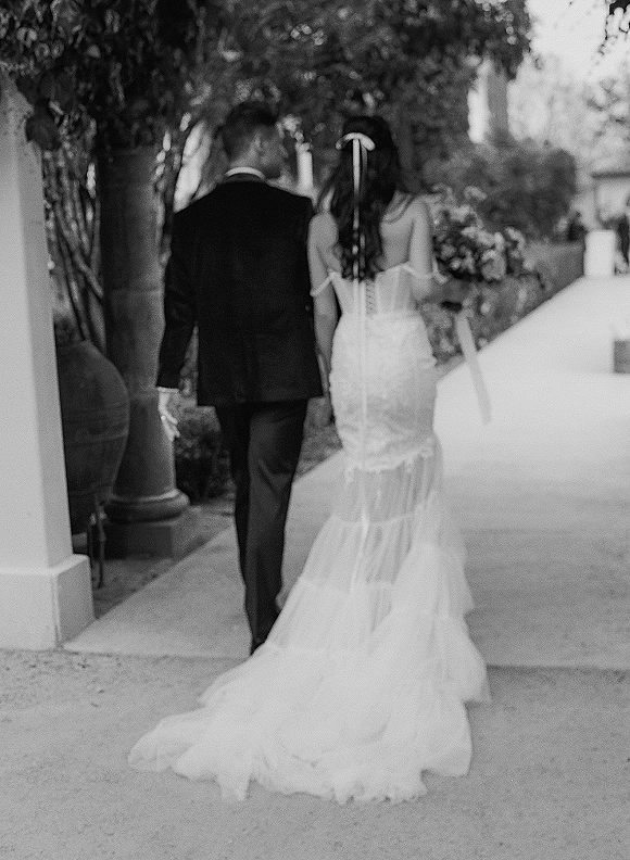 Bride and groom holding hands walk away down a columned outdoor walkway, her dress train and bouquet visible beside his tuxedo