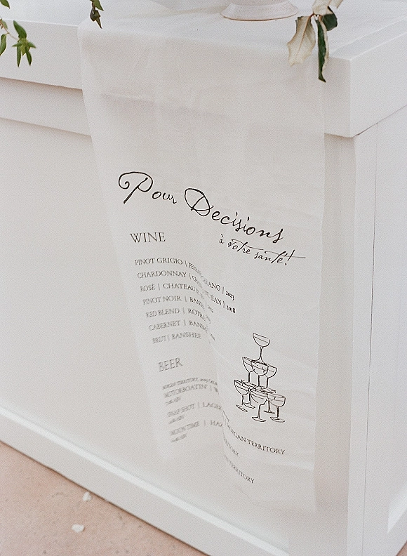 Wedding bar menu fabric sign with calligraphy drink list and champagne coupe illustration, greenery accent on a white counter indoors