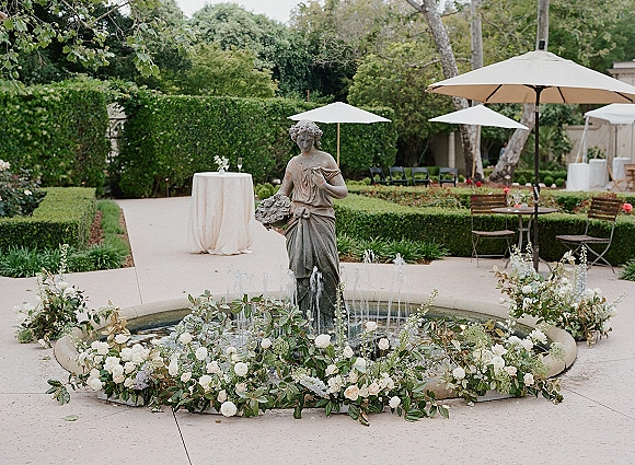 Cocktail hour decor with a wedding cocktail hour setup around a fountain statue, white rose garland, champagne flutes, and patio umbrellas in a hedge garden