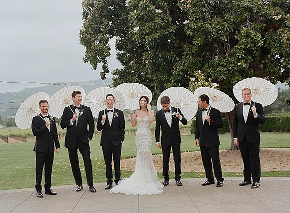 Wedding party portrait of bride in a strapless lace gown with groomsmen in black tuxedos holding white paper parasols under a large tree