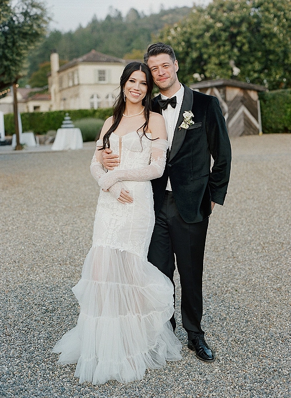 Couple portrait of bride and groom smiling in an embrace, her off-the-shoulder lace dress, in a gravel courtyard by an estate building