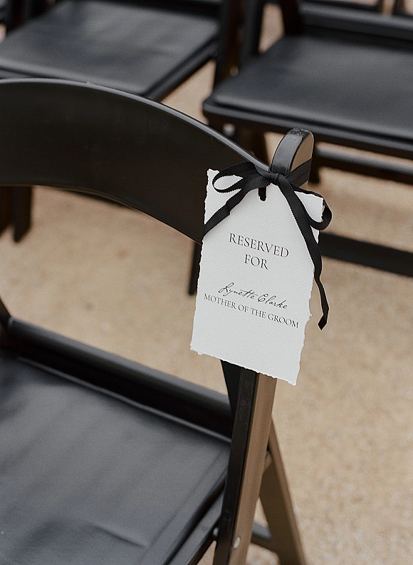 Reserved seat sign with calligraphy tied by a black ribbon bow to a black folding chair in ceremony rows on a stone patio floor