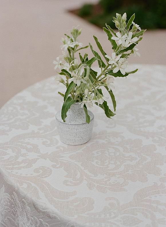 Wedding centerpiece with white flowers and greenery in a small ceramic vase on patterned tablecloth, set on an outdoor patio by hedges