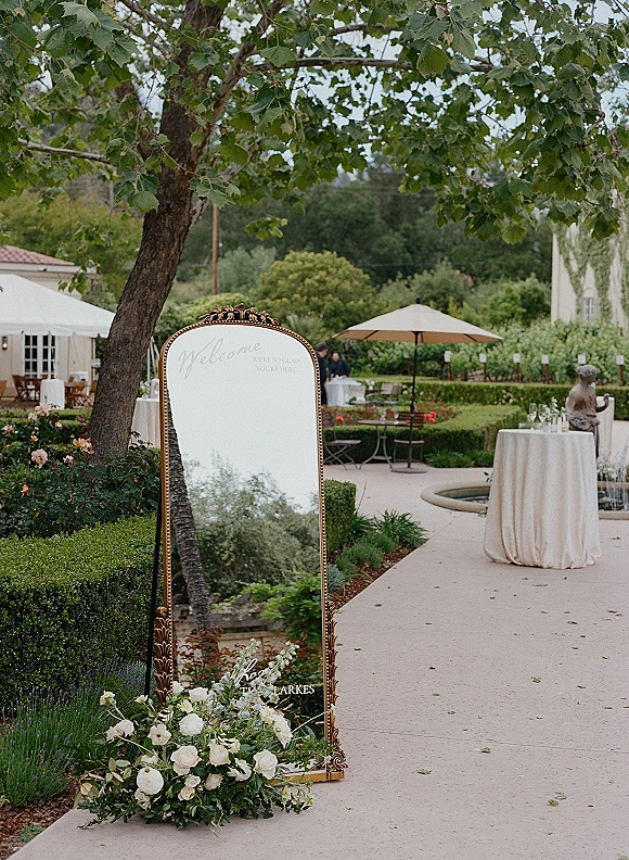 Wedding welcome sign on a gold ornate mirror with calligraphy and white floral arrangement, set in a garden courtyard near a fountain