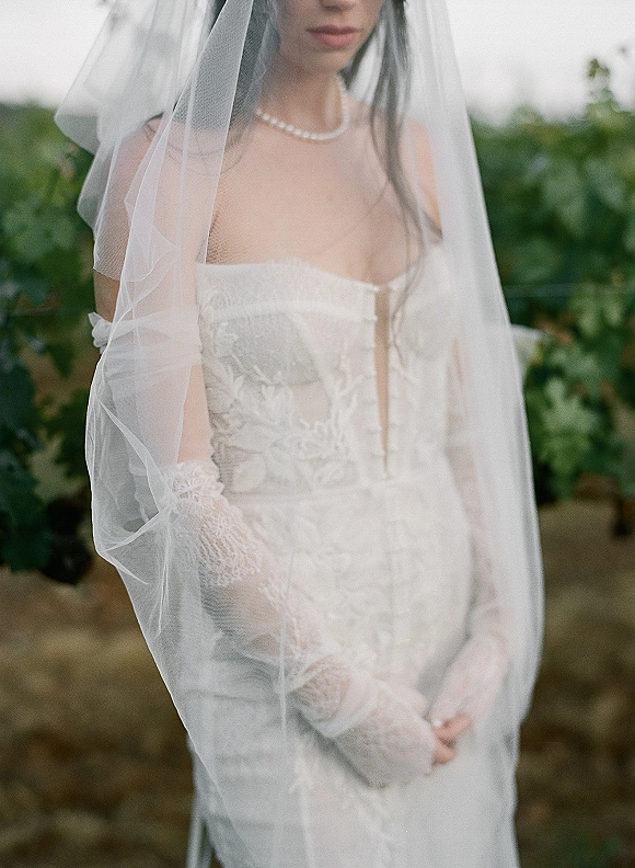 Bridal portrait of a bride with veil wearing a strapless lace wedding dress and pearl necklace on an outdoor path with garden greenery