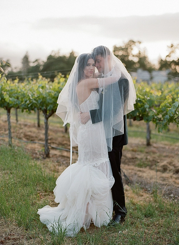 Couple portrait of bride and groom under veil, bride in lace mermaid gown hugging groom in suit beside vineyard rows at sunset