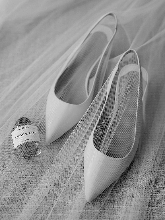 Bridal shoes, white pointed-toe heels arranged with a bridal veil and perfume bottle on a soft fabric surface flat lay