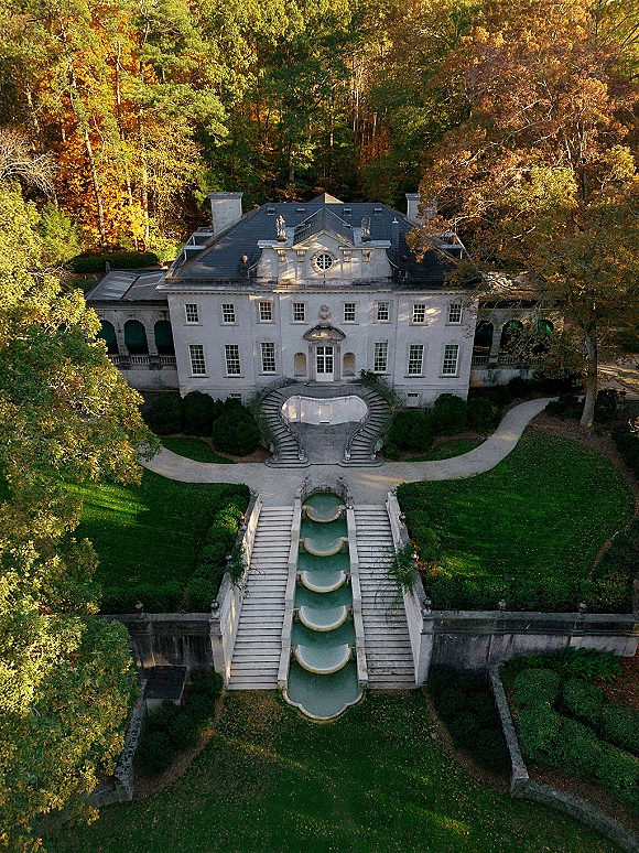 Wedding venue exterior of a stone mansion with a grand staircase and tiered fountain, framed by manicured hedges and autumn trees