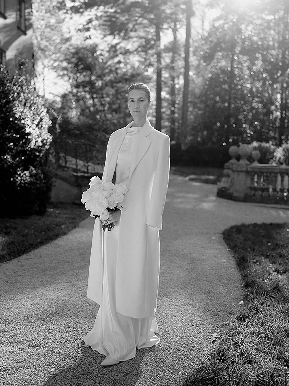 Bridal portrait of a bride holding a bouquet in a black and white bridal portrait, walking a garden path by a stone balustrade and manor exterior
