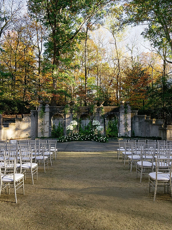 Outdoor ceremony setup with white floral arrangements and greenery garlands framing a stone wall altar, silver chiavari chairs on lawn amid autumn trees