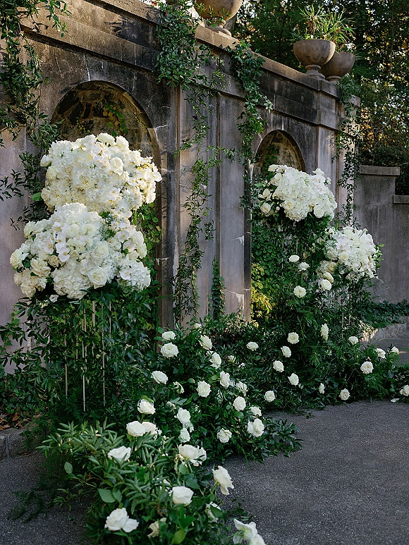 Wedding ceremony florals with white hydrangea wedding flowers lining a paved aisle, framed by floral pillars against a stone wall with arches