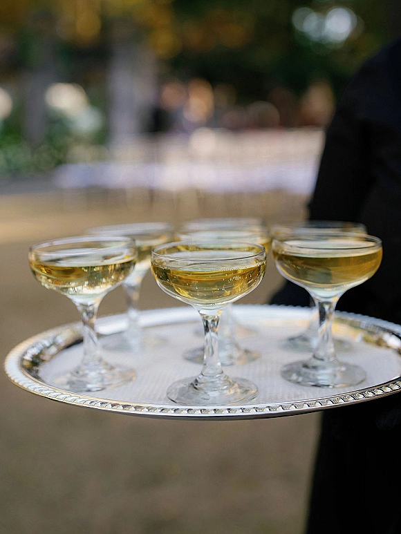 Champagne service with champagne coupe glasses on a silver serving tray, passed along an outdoor walkway with blurred guests and trees