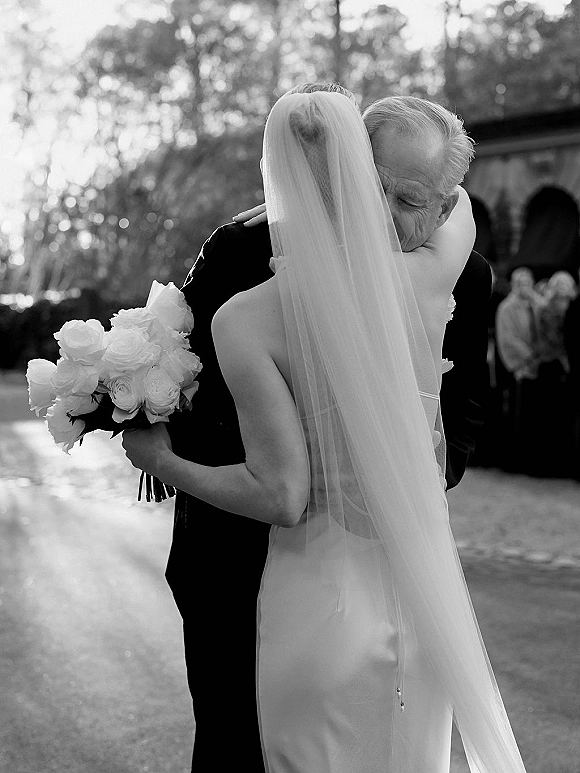 Wedding hug as bride hugs her father, holding a white rose bouquet with a long veil flowing in an outdoor stone courtyard among guests