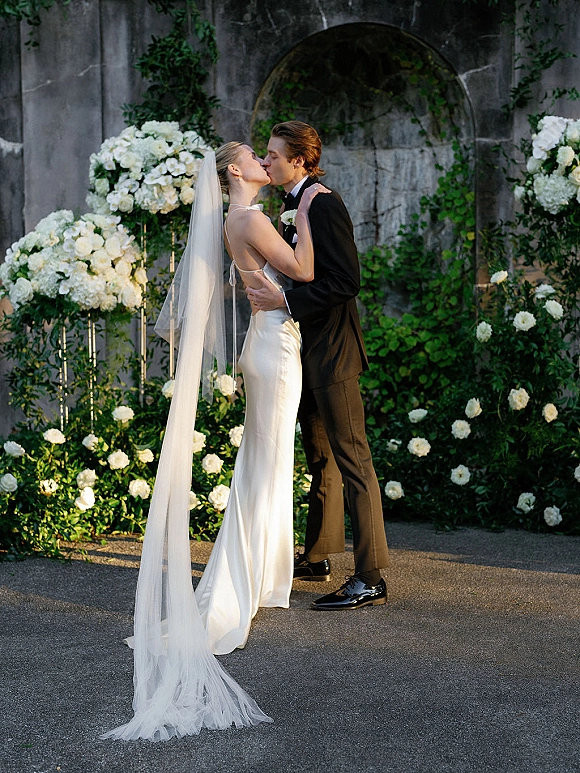 Wedding kiss portrait of bride and groom kissing in satin gown and black tux, veil trailing beside white roses and greenery by stone wall
