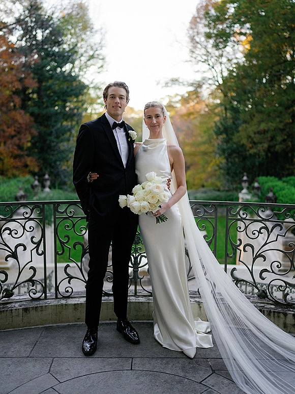 Couple portrait of bride and groom in black-tie attire, her long veil flowing as she holds a white rose bouquet on a garden terrace