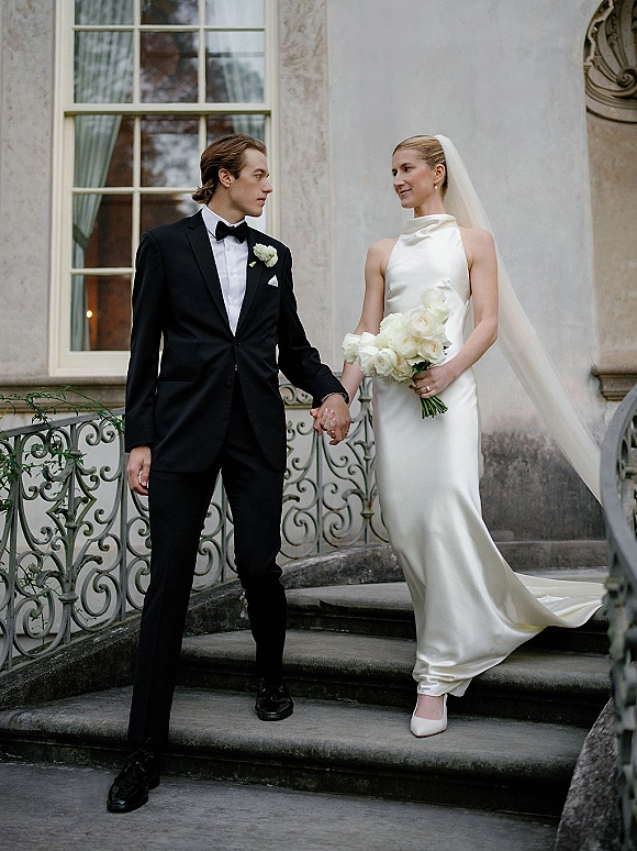 Couple portrait of bride and groom holding hands on a stone staircase, her veil and white rose bouquet beside his black tuxedo bow tie