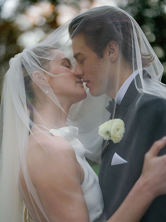Wedding kiss portrait of bride and groom kissing under her veil, strapless gown and tuxedo with boutonniere, framed by trees outdoors