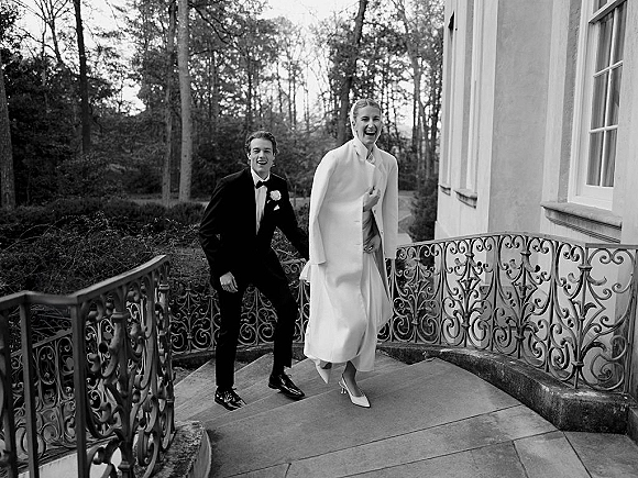 Couple portrait in a black and white wedding photo, bride in a bridal coat and gown beside groom in tuxedo on stone steps by wrought iron railing