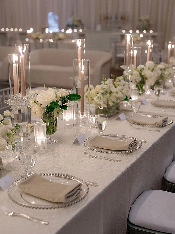 Wedding tablescape with a white wedding centerpiece and glass cylinder candles glowing on ivory linens in a draped reception room near the head table