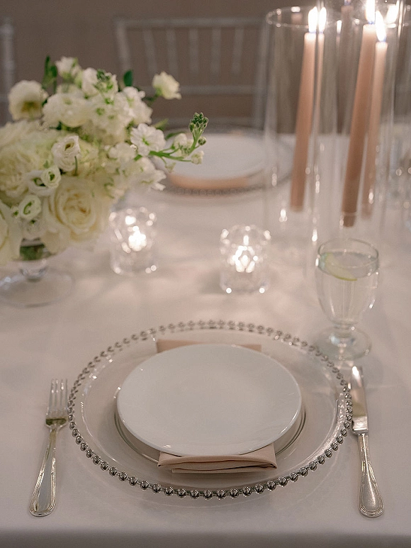 Reception tablescape with wedding place setting, white floral centerpiece and taper candles in glass hurricanes on a white tablecloth with chairs