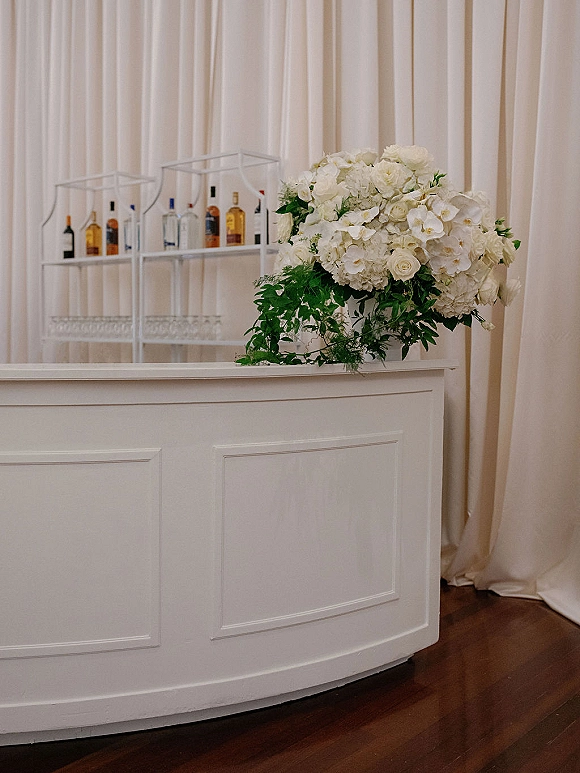 Wedding bar setup with a white wedding bar, liquor bottles and glassware on metal shelves, topped with hydrangea floral accent by white drapes