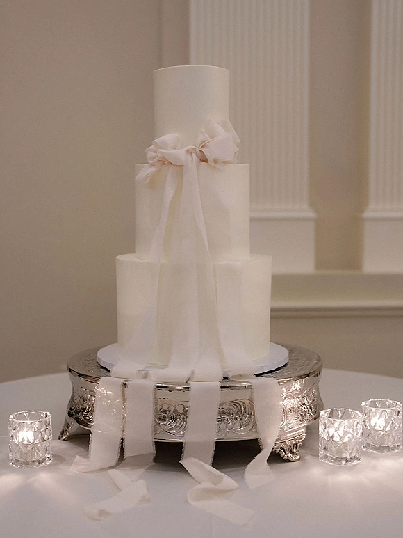 Wedding cake with a white ribbon bow on a silver stand, surrounded by votive candles on a white tablecloth before paneled columns
