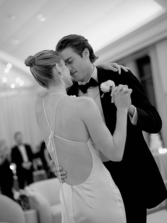 First dance wedding first dance as bride in a wedding dress and groom in tuxedo hold close under ballroom lights with guests blurred behind