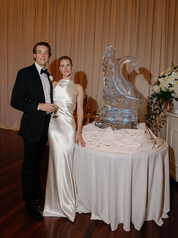 Couple portrait of bride and groom posing in black tie attire beside a swan ice sculpture on a round table with draped curtains behind