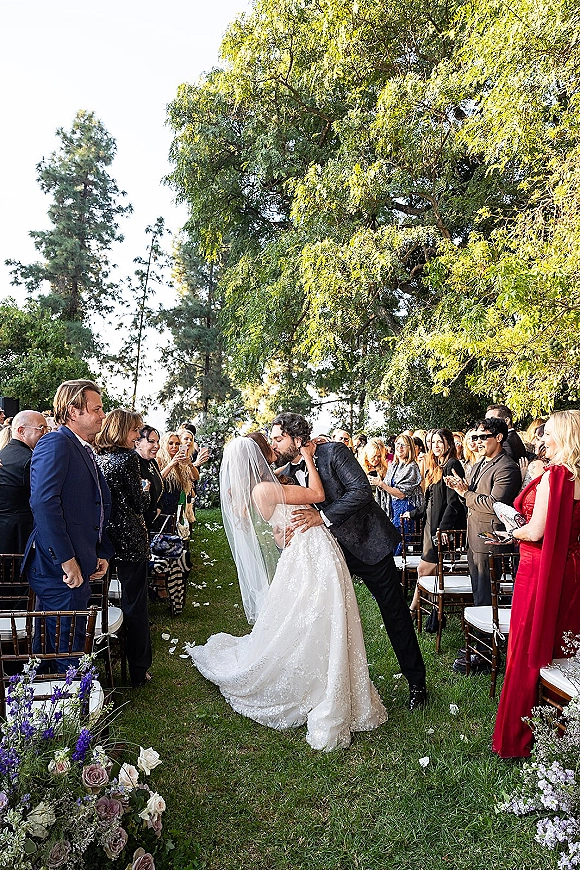 Wedding kiss during a ceremony kiss moment as bride in gown and veil dips with groom in tuxedo on a petal-lined garden aisle