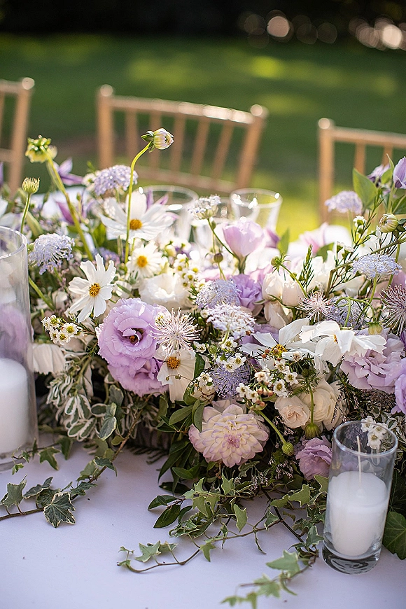 Wedding floral centerpiece with garden wedding centerpiece wildflowers in white and lavender, ivy garland, candles and glassware on a sunlit lawn table