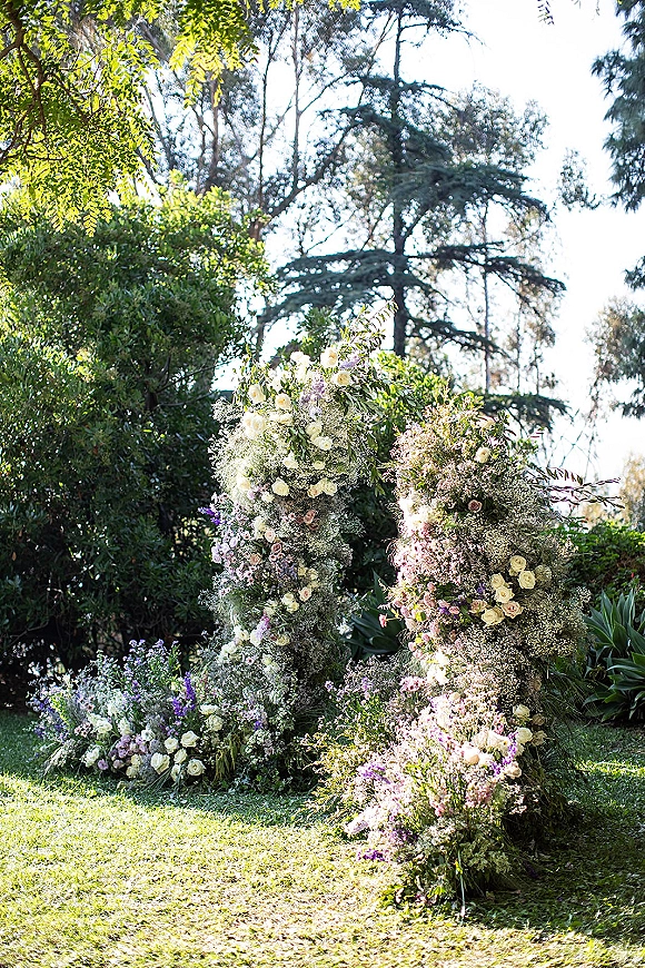 Floral ceremony arch with roses and baby’s breath framed by greenery on a sunlit garden lawn with trees and soft sky backdrop
