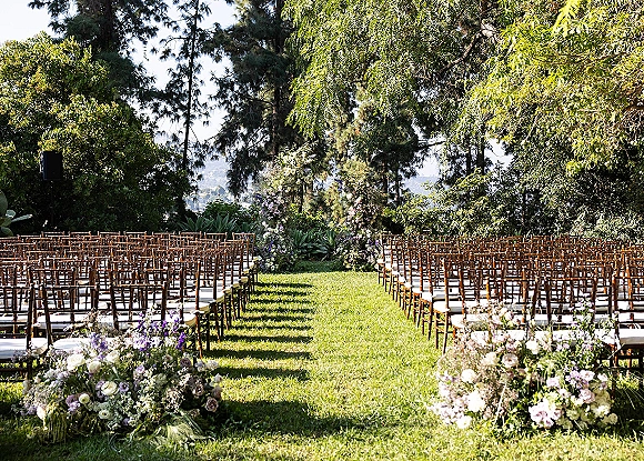 Ceremony setup with wood chairs lining a grass aisle runner, purple and white floral arrangements and greenery on a lawn with hillside view