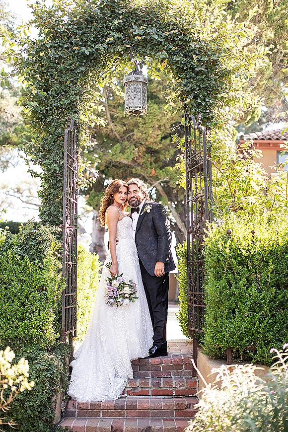 Couple portrait of bride and groom standing under an ivy archway, bride holding bouquet, by a wrought iron gate and brick steps