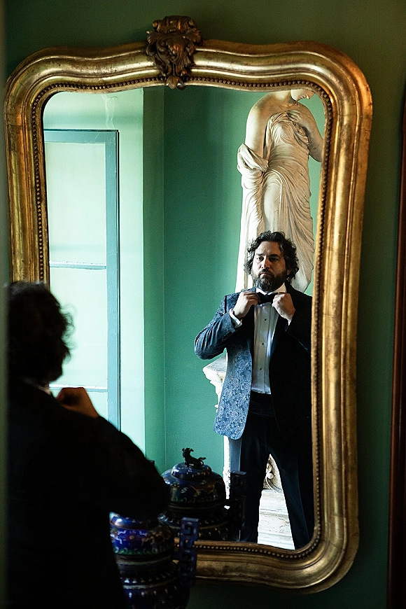 Groom getting ready adjusts his bow tie in an ornate gold mirror, wearing a tuxedo and cufflinks beside a dresser and green wall