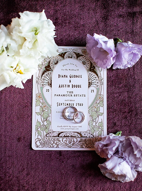 Wedding invitation suite with an art nouveau wedding invitation, rings, and white and lavender flowers on a rich purple velvet backdrop