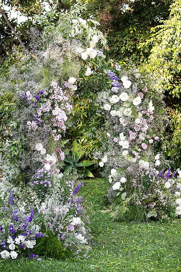Floral ceremony arch with asymmetrical wedding arch design, featuring roses, baby’s breath and greenery on a garden lawn with trees