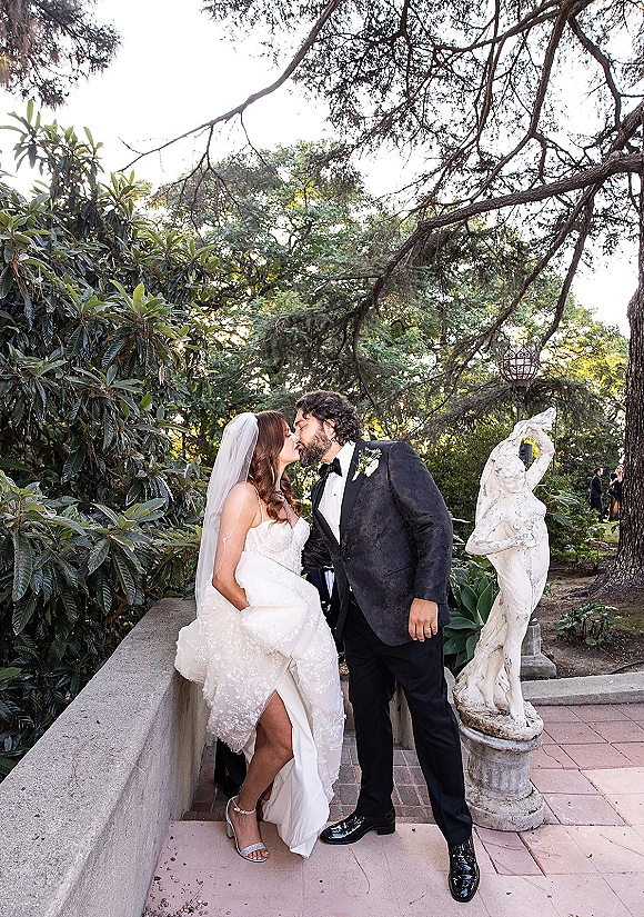 Wedding kiss portrait of bride in strapless lace dress and veil kissing groom in black tux, holding bouquet by stone wall and garden trees