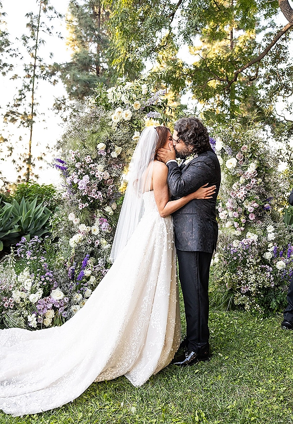 Wedding kiss portrait of bride and groom embracing beneath a floral arch, her cathedral veil and lace train flowing in a garden setting