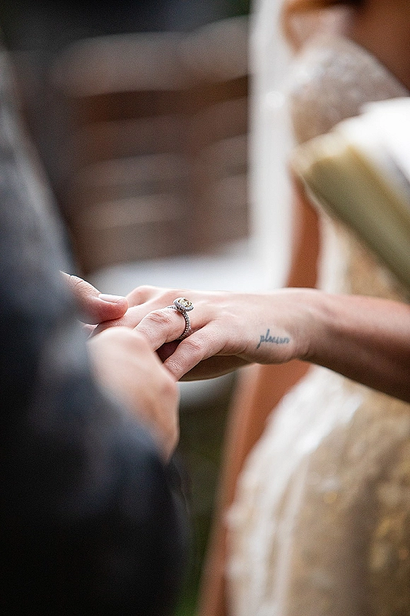 Wedding ring exchange as hands slide a wedding band onto a finger, engagement ring sparkling beside lace wedding dress in an indoor setting
