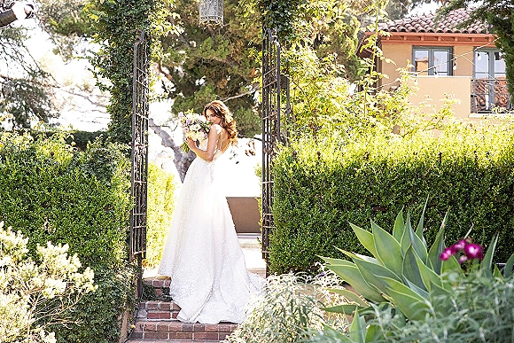 Bridal portrait of a bride holding bouquet, glancing over her shoulder in a strapless gown on brick steps by a greenery arch and wrought iron gate