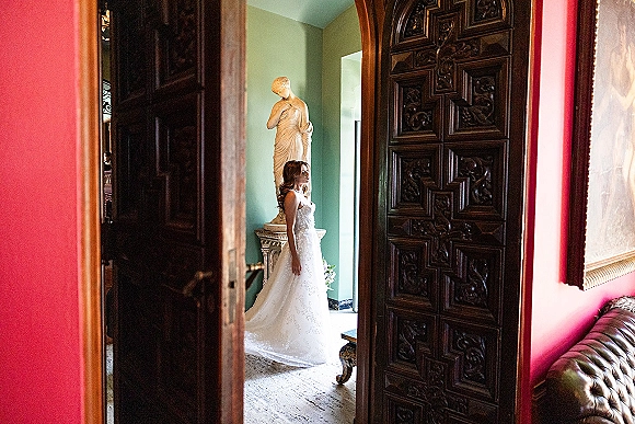 Bridal portrait of a bride standing in doorway, lace gown train and bouquet beside her, lit by window light near carved wooden doors