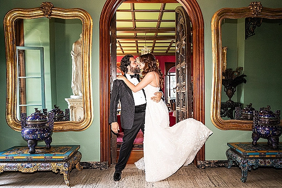 Wedding kiss portrait of bride and groom kissing in a doorway, lace dress train and tuxedo framed by gold mirrors in a green room