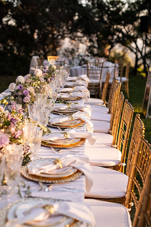Reception tablescape with a floral garland centerpiece on a long banquet table, crystal glassware, gold chargers, and candle accents at sunset lawn reception