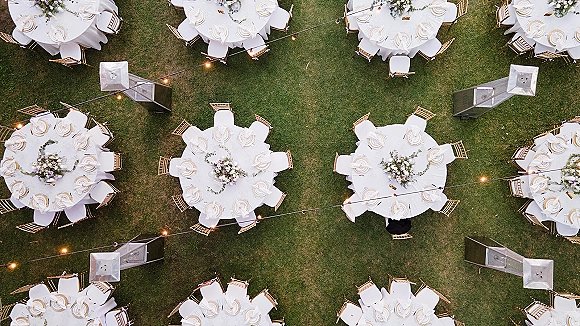 Reception tablescape for an outdoor wedding reception with round tables, white linens, gold chairs, floral centerpieces, and string lights on a grass lawn