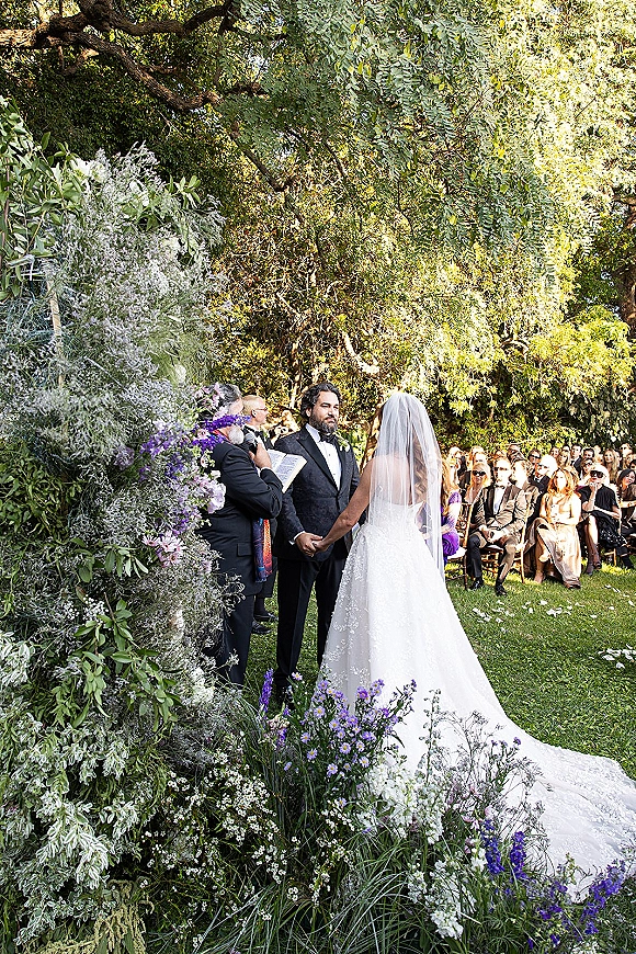 Wedding vows during outdoor wedding ceremony as bride and groom hold hands beneath a floral arch with purple blooms on a garden lawn