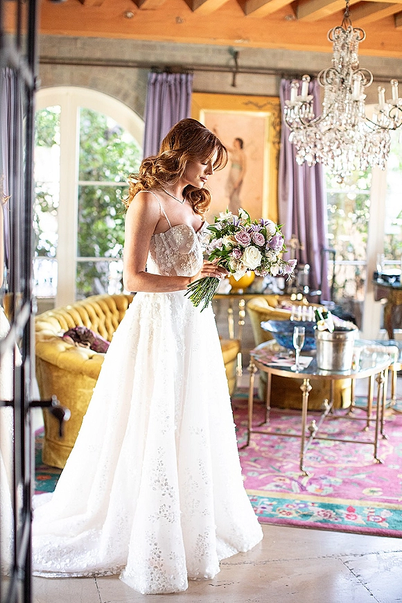 Bridal portrait of a bride holding bouquet of pastel roses in a lace gown, standing by arched windows in an elegant lounge suite