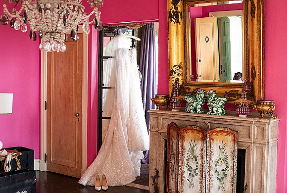 Wedding dress detail with veil hanging above nude heels by a vintage fireplace mantel, framed by an ornate mirror in a pink suite